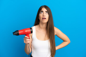 Young caucasian woman holding a hairdryer isolated on blue background looking up and with surprised expression