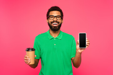 happy african american man in glasses and green polo shirt holding paper cup and smartphone with blank screen isolated on pink