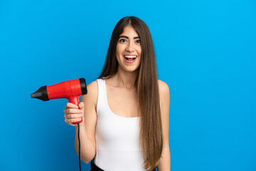 Young caucasian woman holding a hairdryer isolated on blue background with surprise facial...