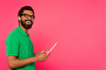 cheerful african american man in glasses and green polo shirt using digital tablet isolated on pink