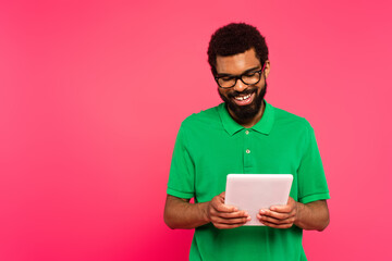 happy african american man in glasses and green polo shirt using digital tablet isolated on pink