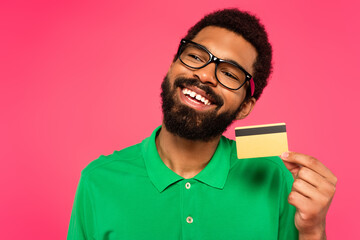 pleased african american man in glasses holding credit card isolated on pink