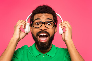 excited african american man in glasses holding wireless headphones isolated on pink