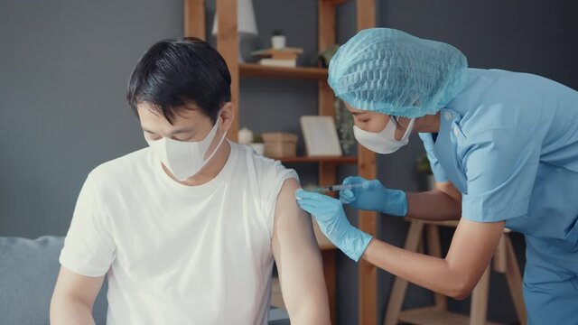 Young Asia lady nurse giving Covid-19 or flu antivirus vaccine shot to male patient wear face mask protection from virus disease sit on couch in living room at house. Vaccination concept.