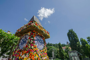 The famous clock tower, one of the city of Graz attractions, made of flowers and the real Clock Tower (Grazer Uhrturm) in the background in Styria region, Austria, in sunny spring day