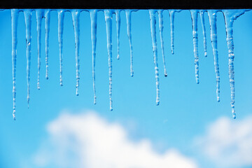Icicles on a blue sky background with clouds. Spring or winter theme with transparent clear thawing ice shining in the sun. Frozen water, water drops, droplets, icicles, thaw.