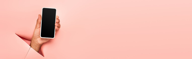 partial view of african american man holding smartphone with blank screen on ripped pink background, banner