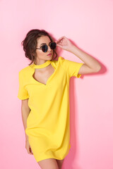Beautiful girl in yellow dress wearing sunglasses posing, smiling on pink background in studio. 
