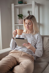Young woman using tablet, sitting on sofa at home, drinking coffee