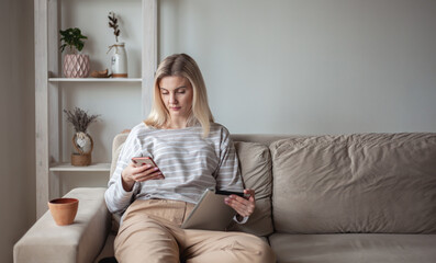 Obraz premium Young woman using tablet and mobile phone, sitting on sofa at ho