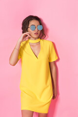 Beautiful girl in yellow dress wearing sunglasses posing, smiling on pink background in studio. 