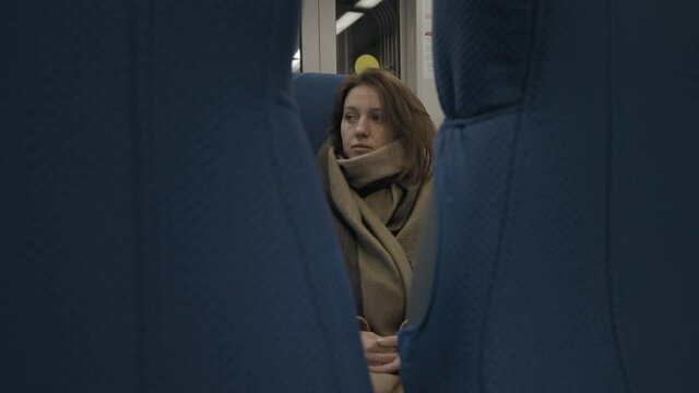 Young woman in beige coat and scarf looking out window while riding train at sunset