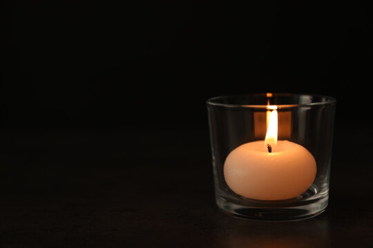 Burning candle in glass holder on table against dark background, space for text