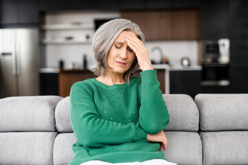 Senior woman with grey hair feel headache, sits on the sofa at home, holds forehead with a pained...