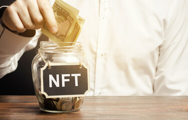 Hand puts dollar bills in glass jar with the word NFT. Non-fungible token. Digitally represented...