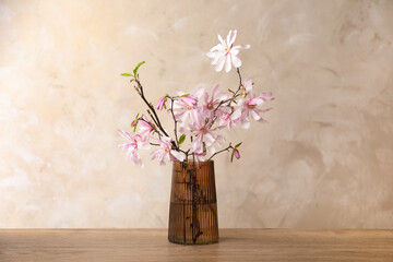 Magnolia tree branches with beautiful flowers in glass vase on wooden table against beige background