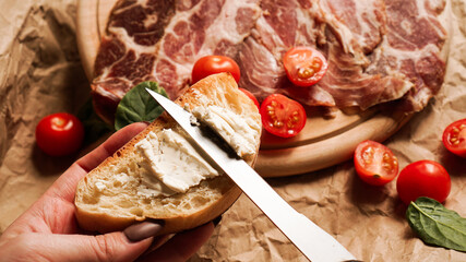 A woman's hand holds a bruschetta and spreads curd cheese with a knife