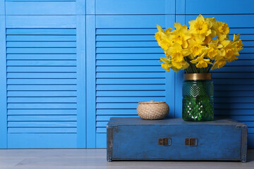 Vase with beautiful daffodils on wooden crate near light blue folding screen indoors, space for text