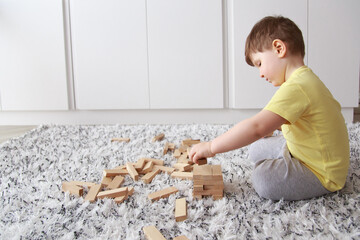 A little caucasian boy in a yellow T-shirt and grey pants sits sideways in a bright room on a fluffy black-and-white carpet and assembles a tower of wooden blocks. 