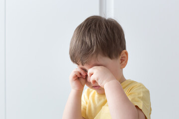 Upset little caucasian boy  in a yellow t-shirt and a large bright room covering his face with his fists and crying