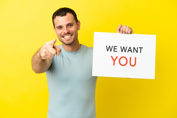 Brazilian man over isolated purple background holding We Want You board and pointing to the front