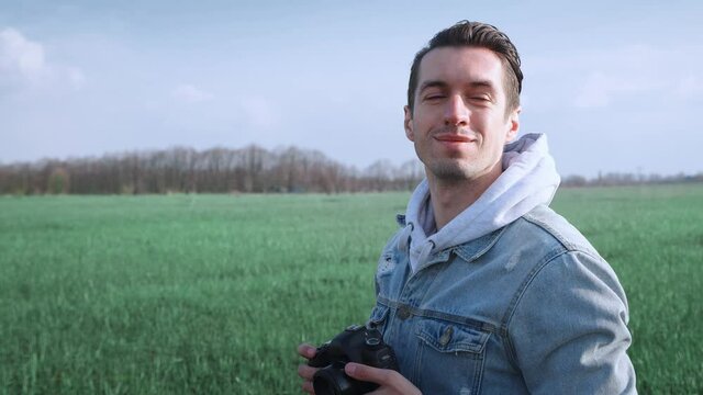 Young male photographer taking photo on professional photocamera, standing at beautiful green field. Portrait of man using digital camera outdoors