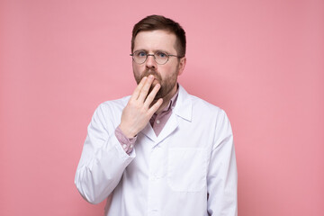Alarmed young doctor in glasses and a white coat covered mouth with hand and looked fearfully into the camera. Pink background.