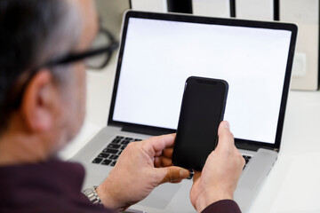 Mani di un uomo tengono uno smartphone e sullo sfondi si vede un computer portatile acceso