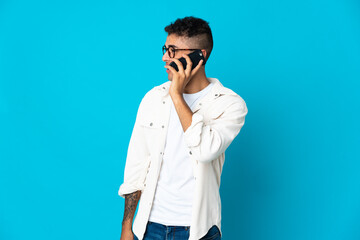 Young Brazilian man isolated on blue background keeping a conversation with the mobile phone with someone