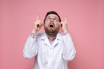 Shocked doctor in a white coat and glasses points index fingers up and looks frightened. Pink...