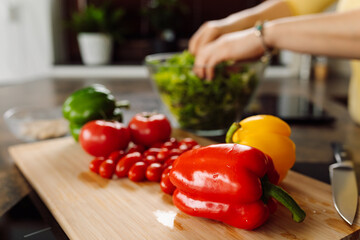 Focus on fresh vegetables in foreground. female hands in background are laying greens in glass salad bowl. Domestic life. healthy vegan food concept