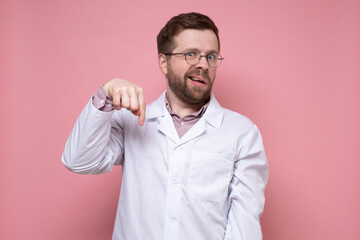 Surprised young doctor points down with index finger and looks puzzled with a strange grimace. Pink background.