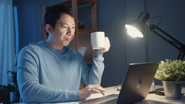 Asia businessman take a break with a cup of coffee relax and checking work assignment at laptop computer for agenda in living room at home overtime at night, Work from home corona pandemic concept.