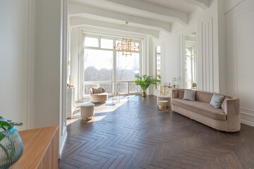 very light and bright interior of luxurious cozy living room with chic soft beige furniture with gold metallic elements, huge window to the floor and wooden parquet