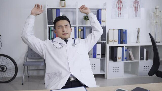 tired asian doctor resting comfortably in chair with hands behind head is closing eyes and exercising neck muscle during break from work at a medical workspace.