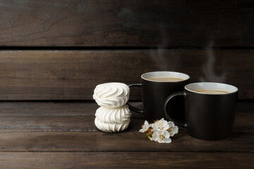 hot coffee and marshmallows on a dark woody background. two black cups of coffee with steam and marshmallows decorated with white flowers.