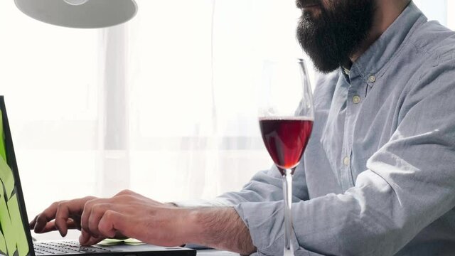 Close up of a bearded man that works at the computer full of reminder notes, man have a glass of wine near laptop to enjoy it.