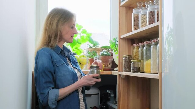 Storing food in pantry, mature female looking at jar of spices holding bowl of green apples