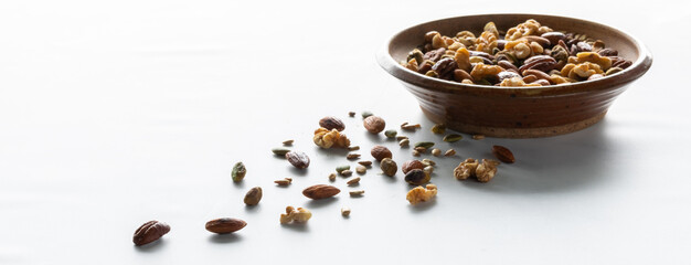 Narrow view of a bowl of trail mix with nuts with several scattered on the table in front, against a white background.