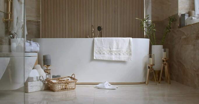 Bathroom tiled with marble tiles in different shades of brown. The plumbing is gold-colored. Baskets with towels is on the floor near big white bathtub. On the left in the frame is a closed toilet bow