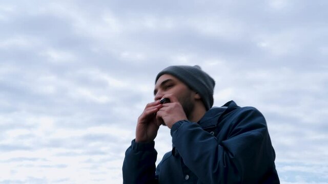 Boy with beard of Caucasian appearance in gray hat sits against the blue sky creates music with his mouth and enjoys life. Young male musician plays the harmonica.