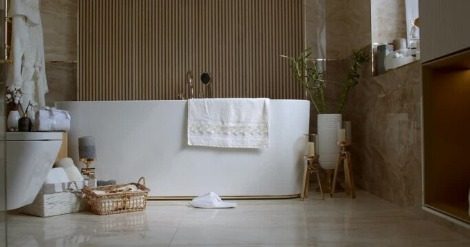 Bathroom tiled with marble tiles in different shades of brown. The plumbing is gold-colored. Baskets with towels is on the floor near big white bathtub. On the left in the frame is a closed toilet bow