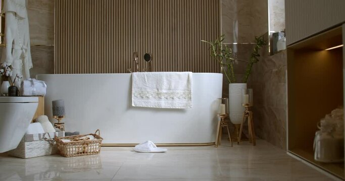 Bathroom tiled with marble tiles in different shades of brown. The plumbing is gold-colored. Baskets with towels is on the floor near big white bathtub. On the left in the frame is a closed toilet bow