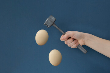 Female hand holds iron hammer ready to hit light brown eggs flying on dark blue background,...