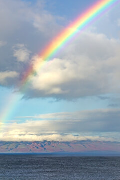 Vibrant and large beautiful rainbow arcing through puffy clouds over the blue ocean with volcano on horizon in Maui, Hawaii on a bright blue sunny day.