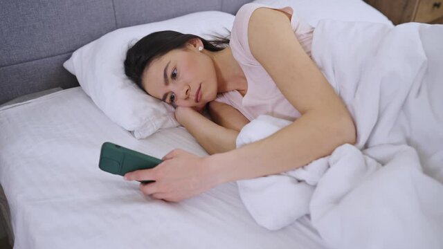 Side view of Pretty young woman lying in bed using mobile phone at home. Waking up with a cellphone concept