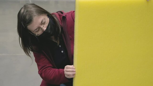 Woman in mask against virus is located in building materials store chooses yellow, soundproof insulation for house to buy. Concept of women independence, gender equality, repair and construction