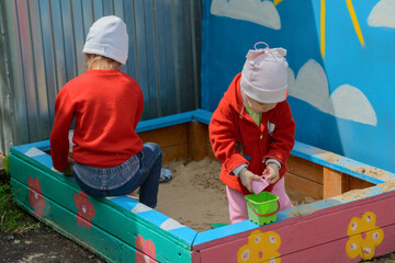 Fototapeta na wymiar Two little girls in red sweatshirts play in the sandbox