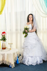 Fototapeta premium A happy Jewish bride stands in the hall before the chuppa ceremony at a table with flowers with a bouquet of white roses in her hands.
