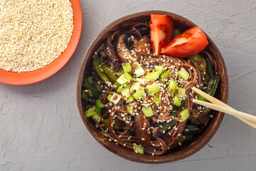 Soba with mushrooms and sesame seeds in a bowl of coconut shells on a concrete background next to sesame seeds and soy sauce and sticks.